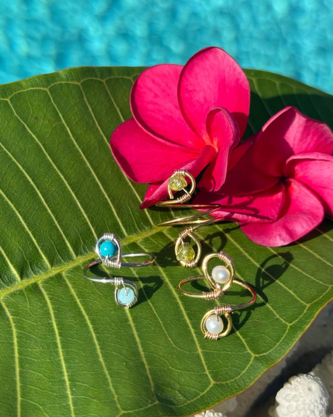 Three rings with gemstones on a green leaf with pink flowers and a pool in the background